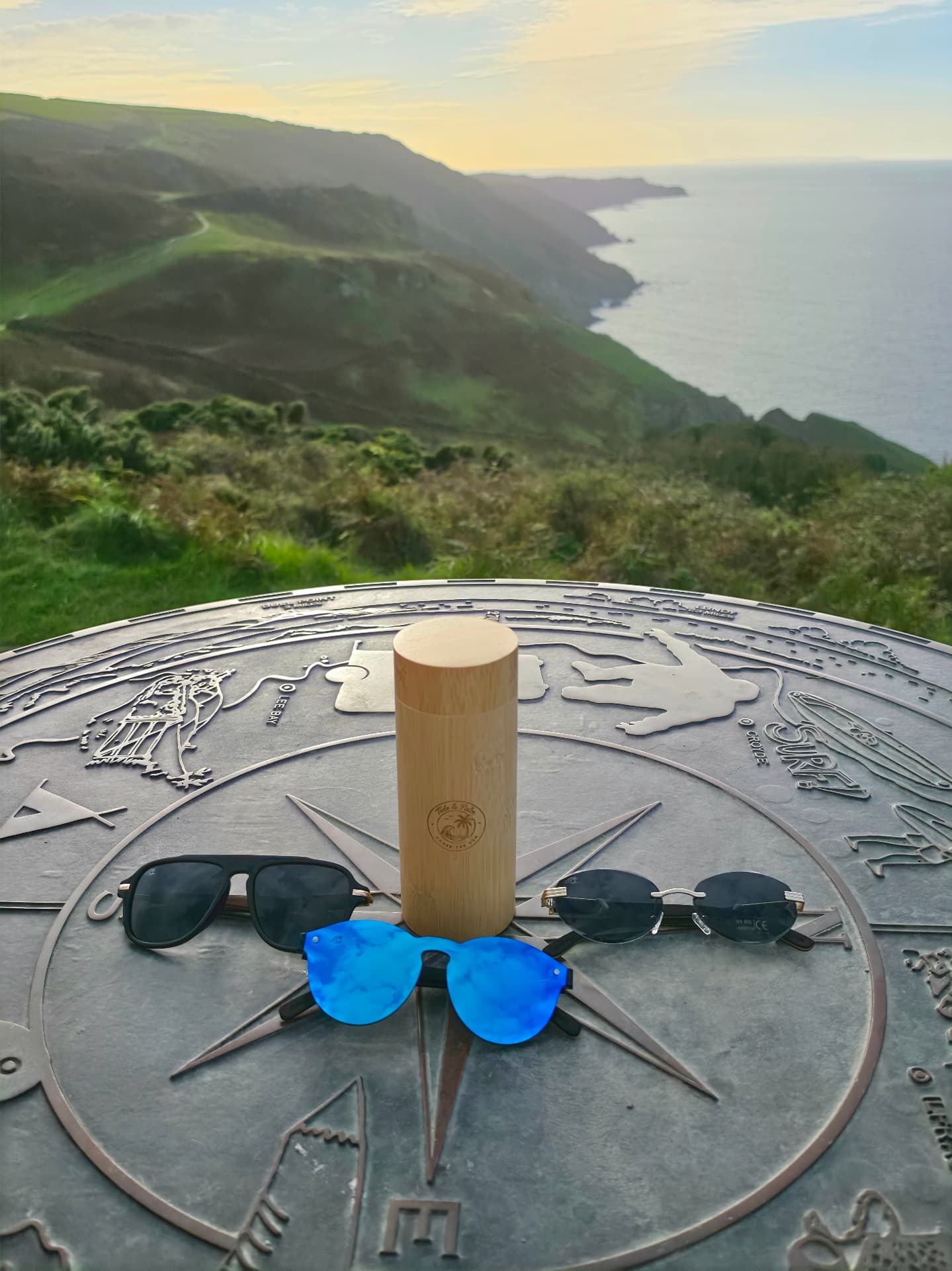The engraved Tide & Palm bamboo hard case with sunglasses on a compass table overlooking the Devon coast