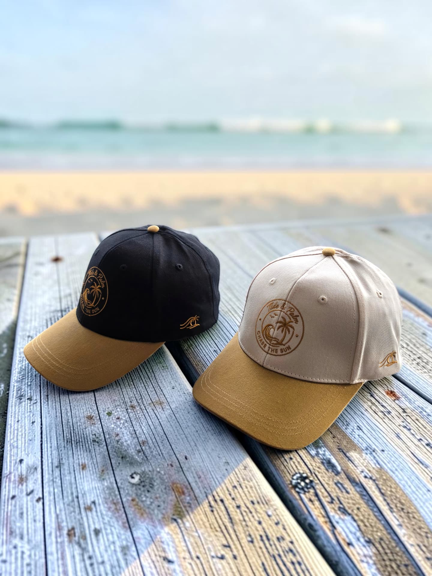 Black and Cream Tide & Palm Chase the Sun caps on a weathered boardwalk above the beach